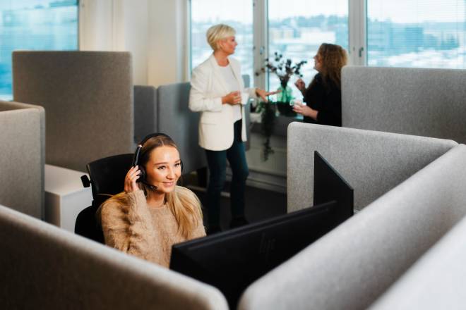 Woman sitting with headphones on in office corner, and 2 other women standing behind her, in front of a window, talking