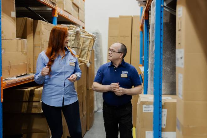One woman in a JYSK shirt and one man in a JYSK polo walk together through a JYSK logistics storage, which part of a JYSK store. Around the two are standing a lot of boxes of new products
