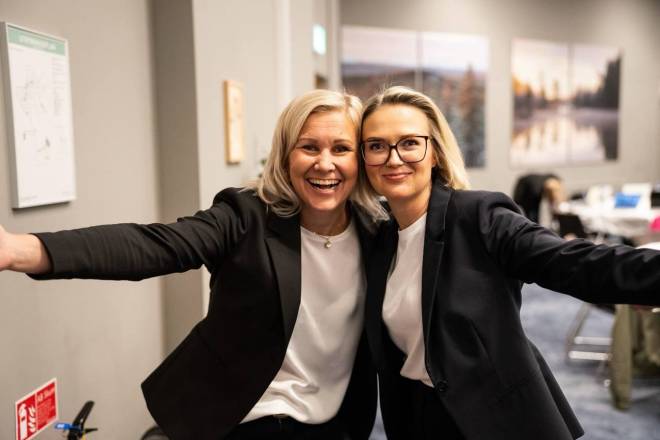 Two blonde women hugging each other half and opening up their bodies and arms towards the camera. One of the women is wearing glasses. Both are wearing black pants, a black blazer and a white t-shirt. In the background you can see an office setting
