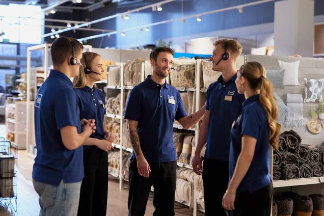 Group of young people (2 girls and 3 boys) is standing in a half circle in a JYSK store in front of textile products, having a team meeting